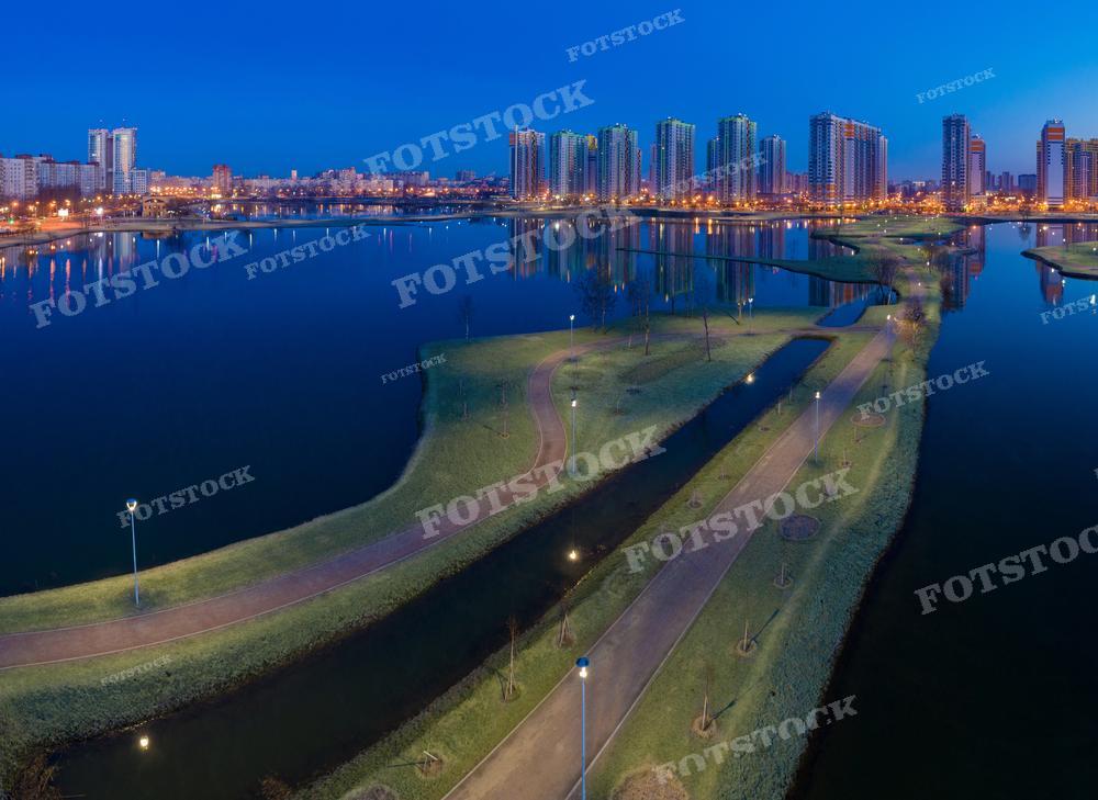 На фотографии запечатлен вечерний городской пейзаж, вид на спальный район города с высоты птичьего полета. На переднем плане видны озера с островами и парком, усеянными дорожками и фонарными столбами. На заднем плане расположены многоэтажные жилые дома с ярко освещенными окнами, отражающимися в водной глади. Фотография сделана в синий час — период сумерек, когда небо принимает насыщенный синий оттенок, что придает изображению особый колорит. Такие снимки хорошо подходят для иллюстрации городской жизни в вечернее время, для туристических материалов и обзоров недвижимости.